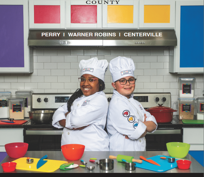 a magazine cover reads At Home in Houston county 2024. Two kids in chefs uniforms standing back to back in front of colorful kitchen background