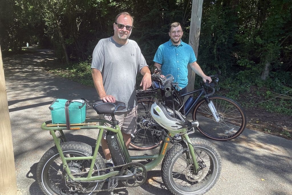 Two men standing at an outdoor trail, one middle-aged, one young adult. Both standing next to bicycles.