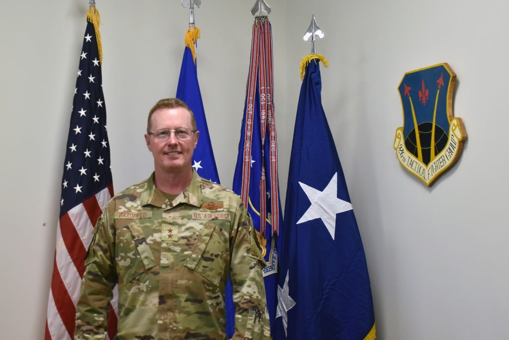 A man wearing glasses and a military uniform, standing in front of various flags