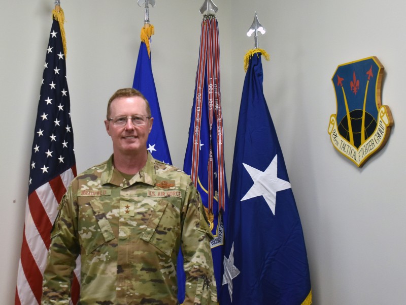 A man wearing glasses and a military uniform, standing in front of various flags