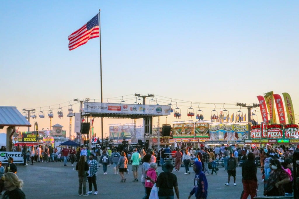 Photo of people walking in front of stage. Sunset and American flag waving in the background.