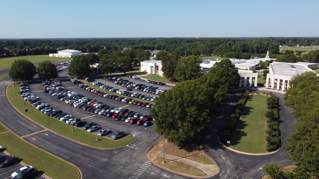aerial view of a parking lot filled with cars, also shows trees and a white building in the photo as well 