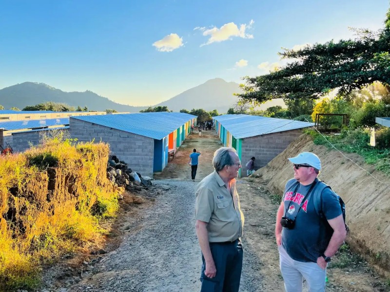 Two men are standing on a dirt path between rows of single-story buildings with colorful doors, under a clear blue sky. One man wears a beige shirt and pants, the other a t-shirt, shorts, and a white cap. In the background, a person walks away from the camera, and a mountain range is visible.