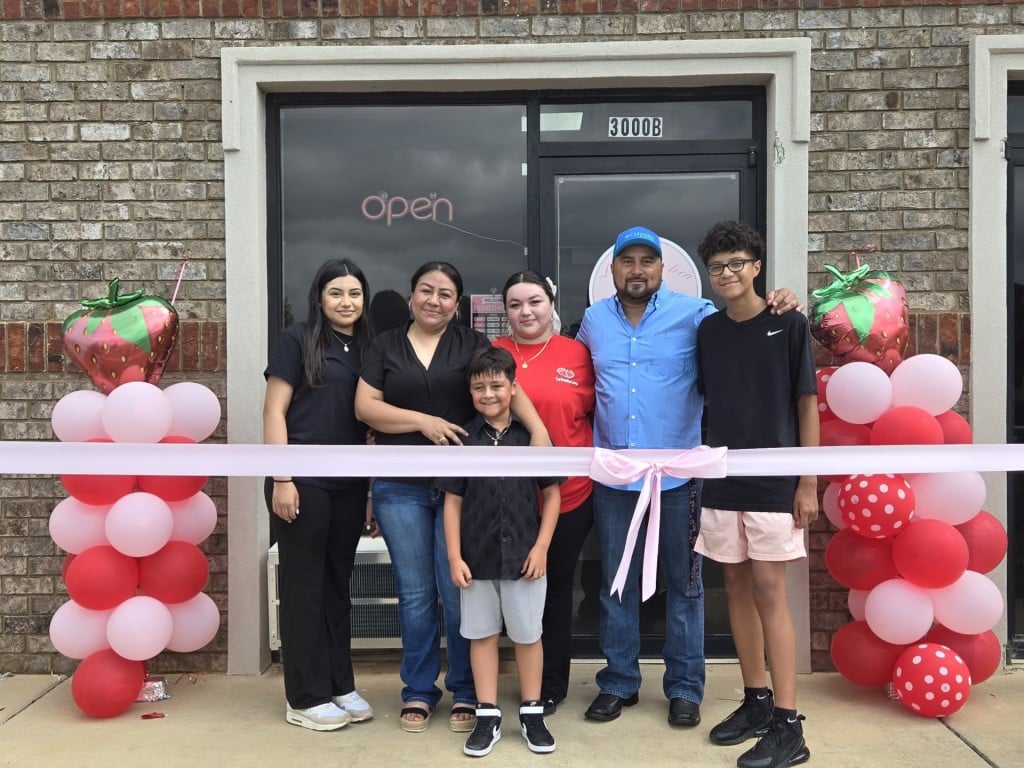 A family smiling at the camera. There are red and pink balloons and a pink ribbon.