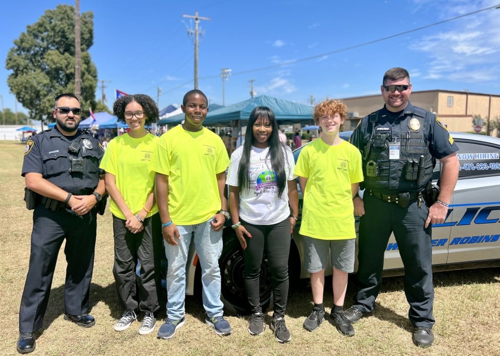 A group photo with the cadets in neon yellow shirts. Two men with police uniforms are also standing with them. Mayor LaRhonda Patrick stands in the middle with a white shirt.