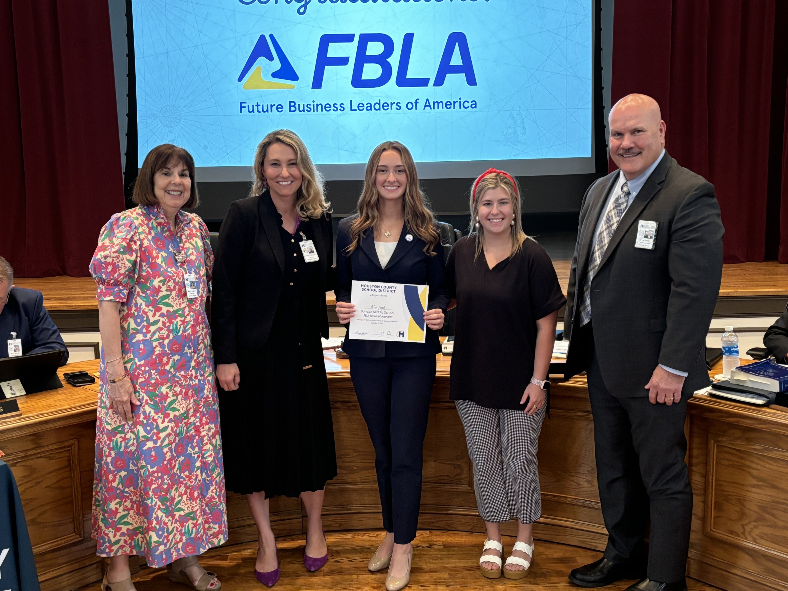 four females, one male posing for a picture, girl in the middle holding up a certificate