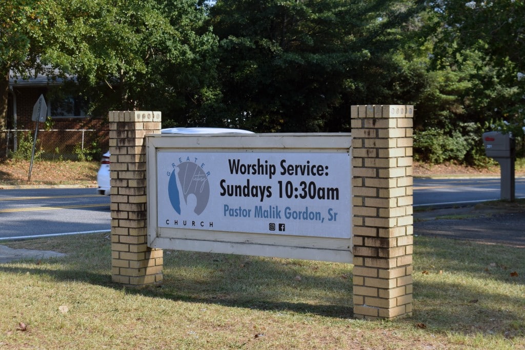 A white sign with black and blue letters. The black letters say "worship service: Sundays 10:30 a.m. The blue letters say "Pastor Malik Gordon, Sr."