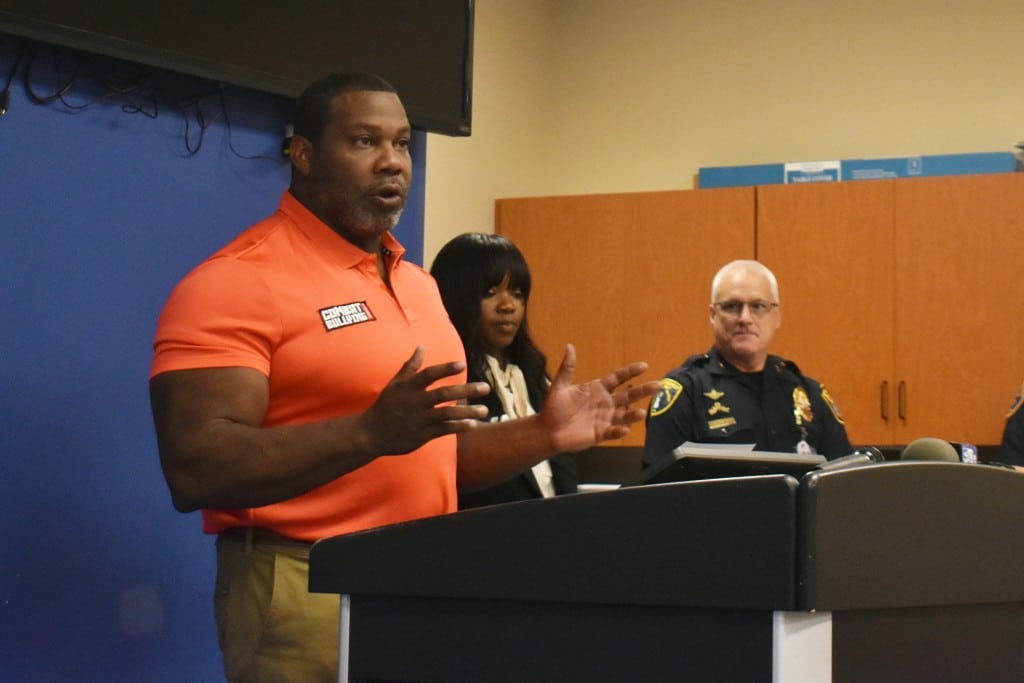A man wearing orange standing behind podium speaking. A woman and a man in police uniform are standing in the background.