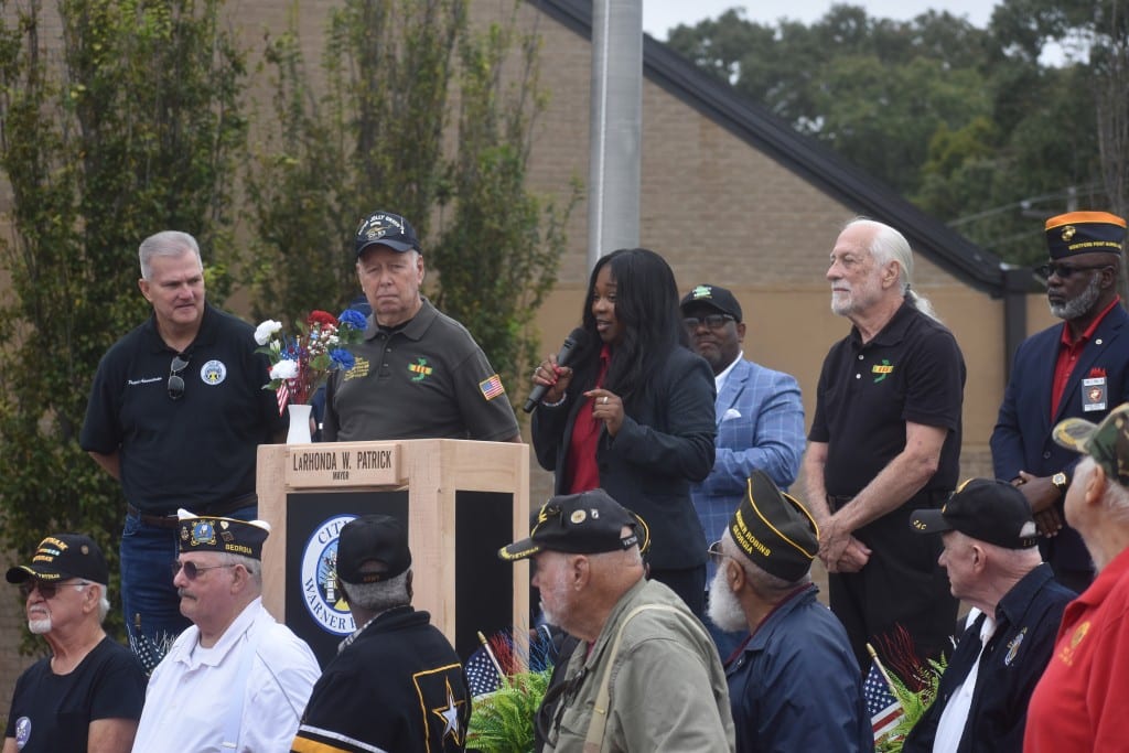 A woman on a podium speaking to the audience. A group of men surround her.