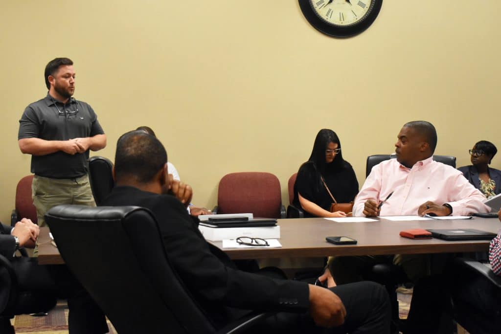 A group of people are in a meeting room. A man standing on the left is speaking to seated attendees around a conference table, which is covered with papers, laptops, and a phone. A wall clock hangs on a beige wall above the group. Most people are looking at the speaker, while one woman with glasses is looking down.