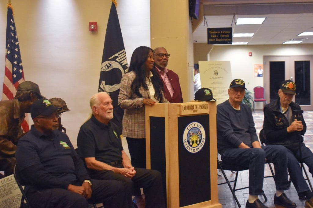 A woman speaking behind a podium. Older men sitting on chairs around her.