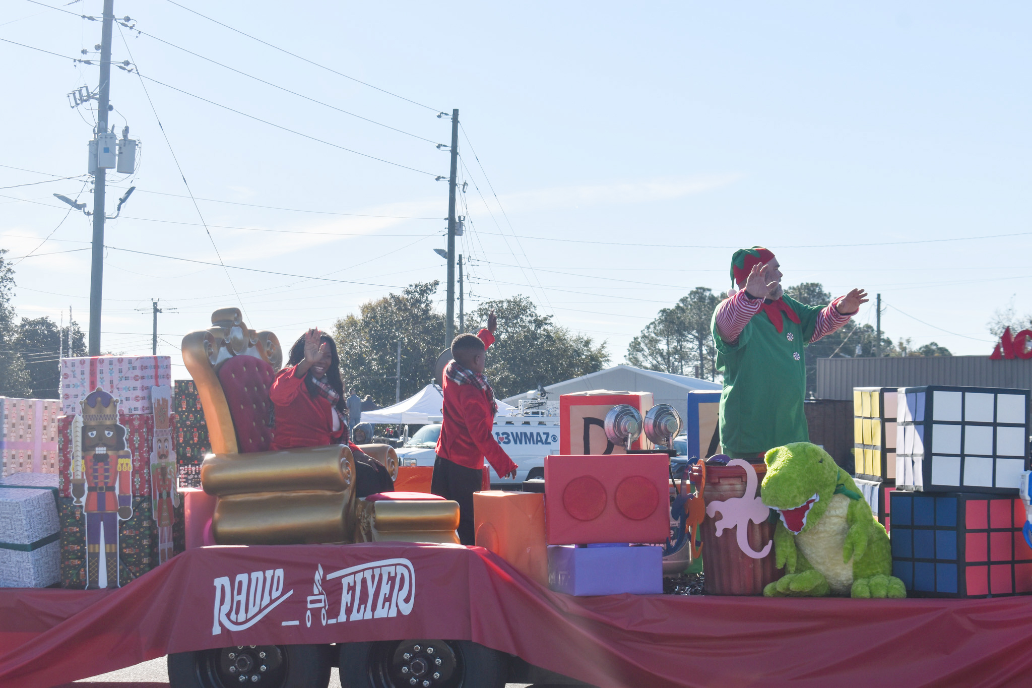 A woman sitting on a throne on a Toyland parade float. A little boy and a man in an elf costume are beside her.