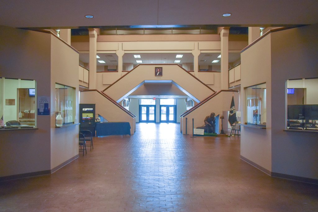 A spacious interior of a building with symmetrical architecture and a central staircase leading to an upper level. The walls are beige, and the floor is tiled in a reddish-brown color. On the left and right, there are office windows with glass partitions. At the back, double glass doors provide natural light, and a statue is visible near the right staircase.