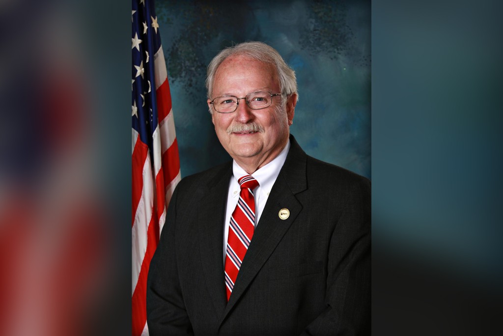 A man in a suit smiling. An American flag is seen in the background.
