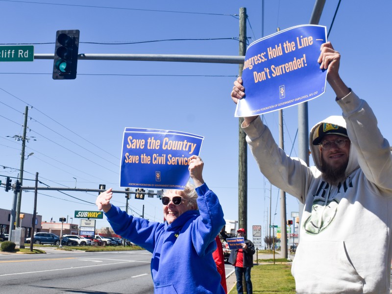 A woman in a blue sweater smiling and waving a sign. A man stands next to her also holding up a sign.