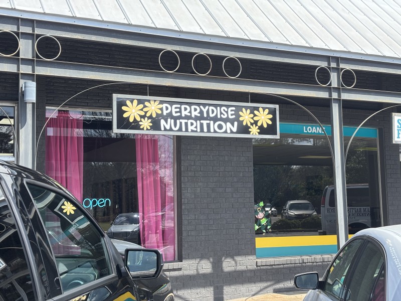 A sign with Perrydise Nutrition written in white letters. Yellow and pink flowers are on the sign.