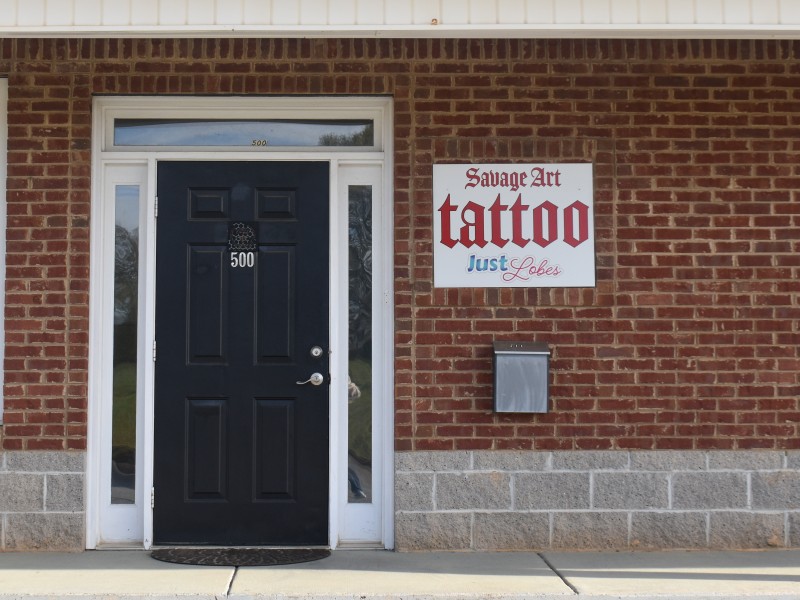 A bricked building with a black door. Next to the door is a sign saying "Savage Art Tattoo" in red letters.