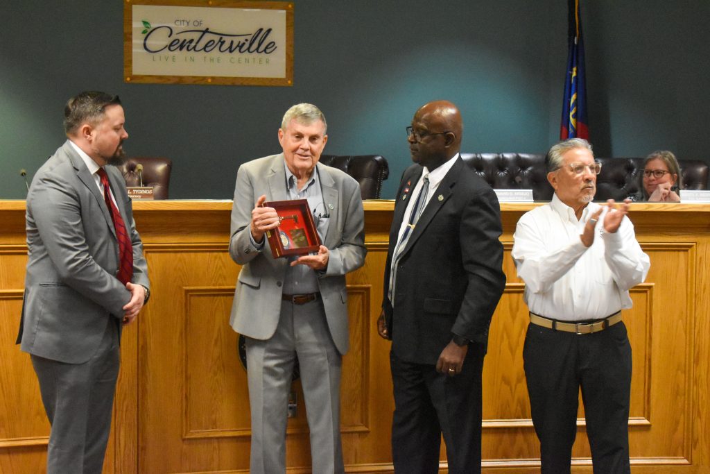 A group of five people is gathered in a formal meeting room with a wooden council desk and a “City of Centerville” sign on the wall. The central figure, an older man in a light gray suit, is holding a framed award with a red background and a gold key inside, smiling as he recieves it. To his right, a man in a dark suit and tie stands attentively. To his left, a man in a white shirt and black pants claps, while another man in a gray suit with a red tie looks on. In the background, a woman wearing glasses sits behind the desk, observing the moment. The setting suggests an official recognition or award ceremony.