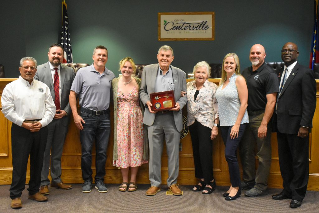 A group of nine people poses in a formal meeting room with a “City of Centerville” sign and an American flag in the background. The central figure, an older man in a light gray suit, holds a framed award featuring a gold key and a medallion. He is surrounded by and officials, all smiling. Some individuals wear business attire, while others are dressed casually. The setting suggests an official recognition or award ceremony, with a mix of personal and professional attendees.