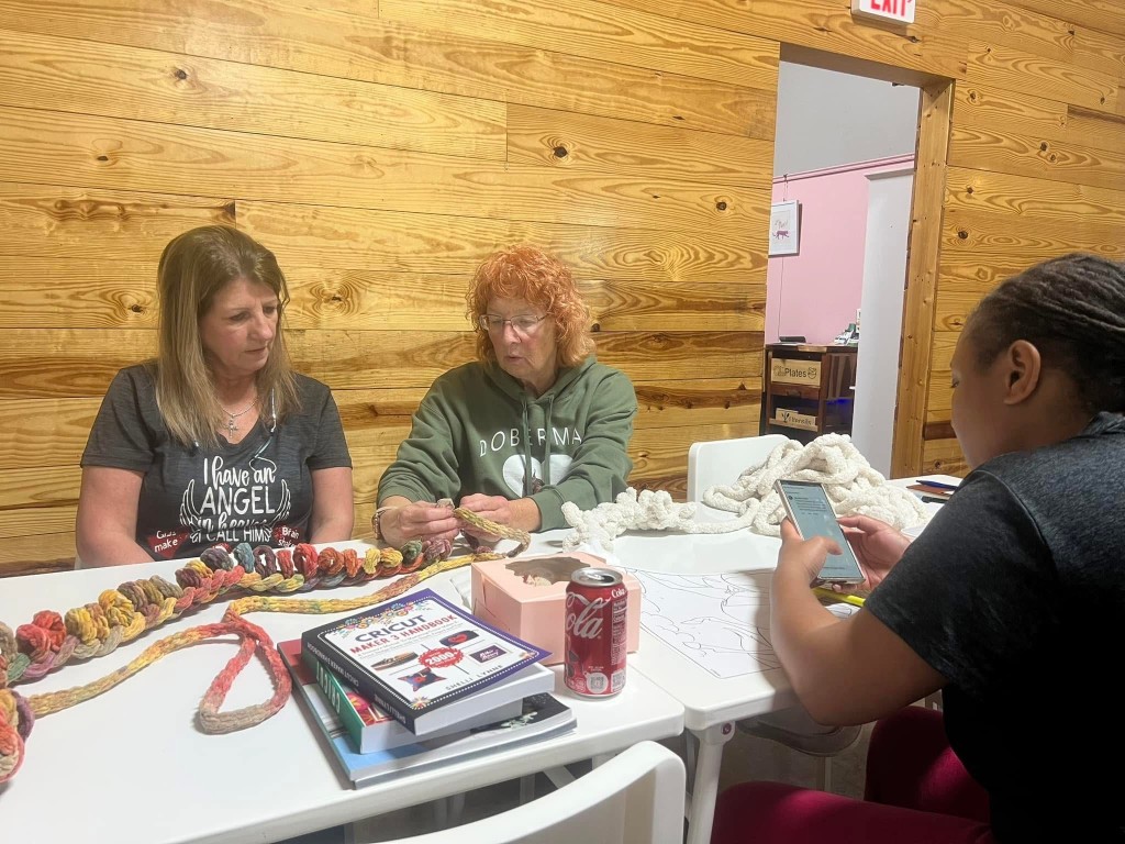 Two women sitting at a table. One of them is wearing a green hoodie and she is teaching a women next to her a blanket.