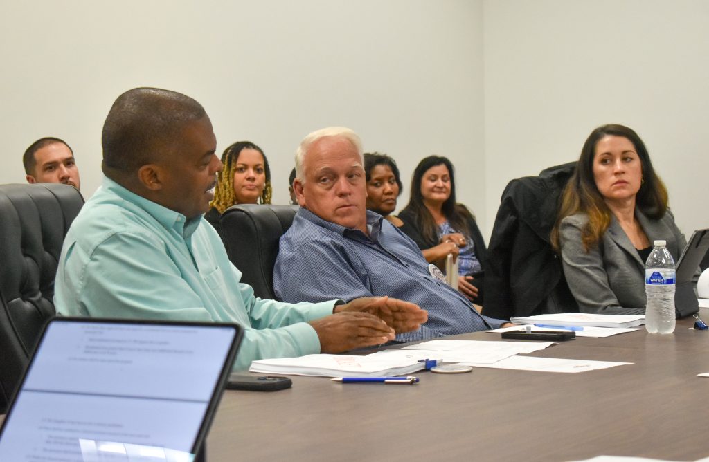 A group of people seated around a conference table engaged in a discussion. A man in a light green shirt is speaking while gesturing with his hands, as others listen attentively. Papers, pens, a water bottle, and laptops are visible on the table. The setting appears to be a professional meeting or workshop in a plain room with white walls.