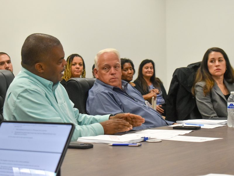 A group of people seated around a conference table engaged in a discussion. A man in a light green shirt is speaking while gesturing with his hands, as others listen attentively. Papers, pens, a water bottle, and laptops are visible on the table. The setting appears to be a professional meeting or workshop in a plain room with white walls.