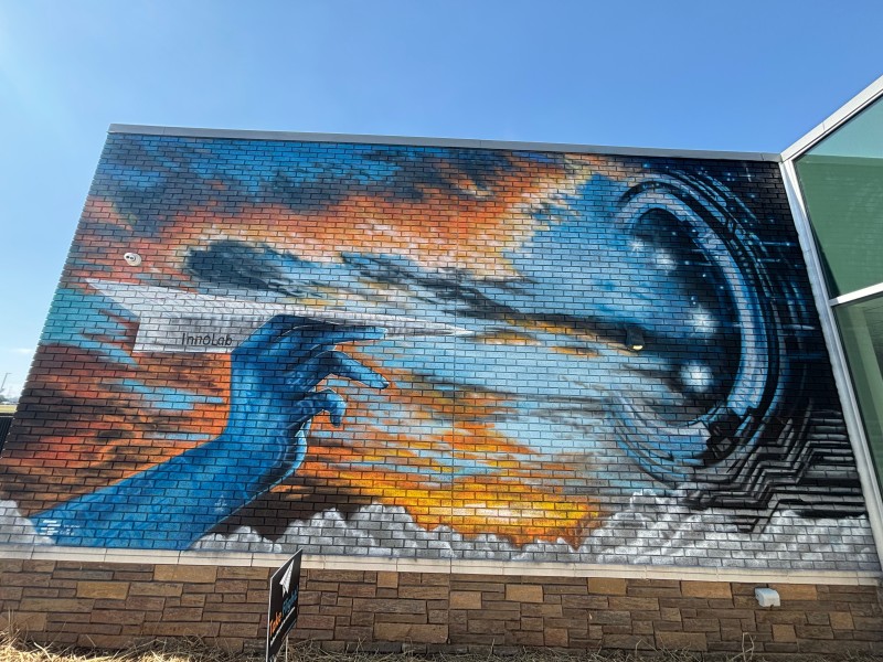 A mural of a blue hand holding a paper airplane. The paper airplane is facing a blue and black portal. Orange is also seen in the background.