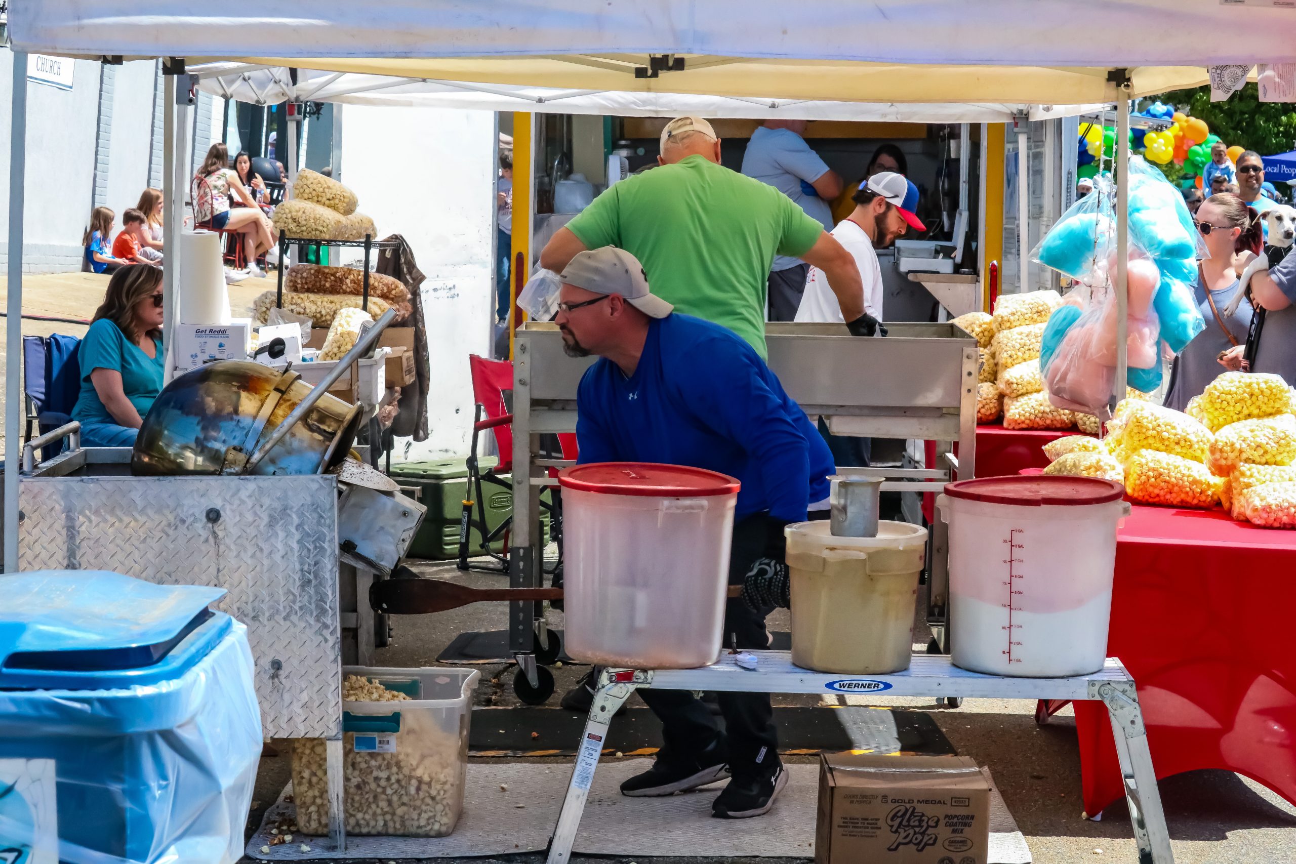 Vendors prepare and package popcorn at an outdoor booth, with bags of popcorn and cotton candy displayed on a red table.