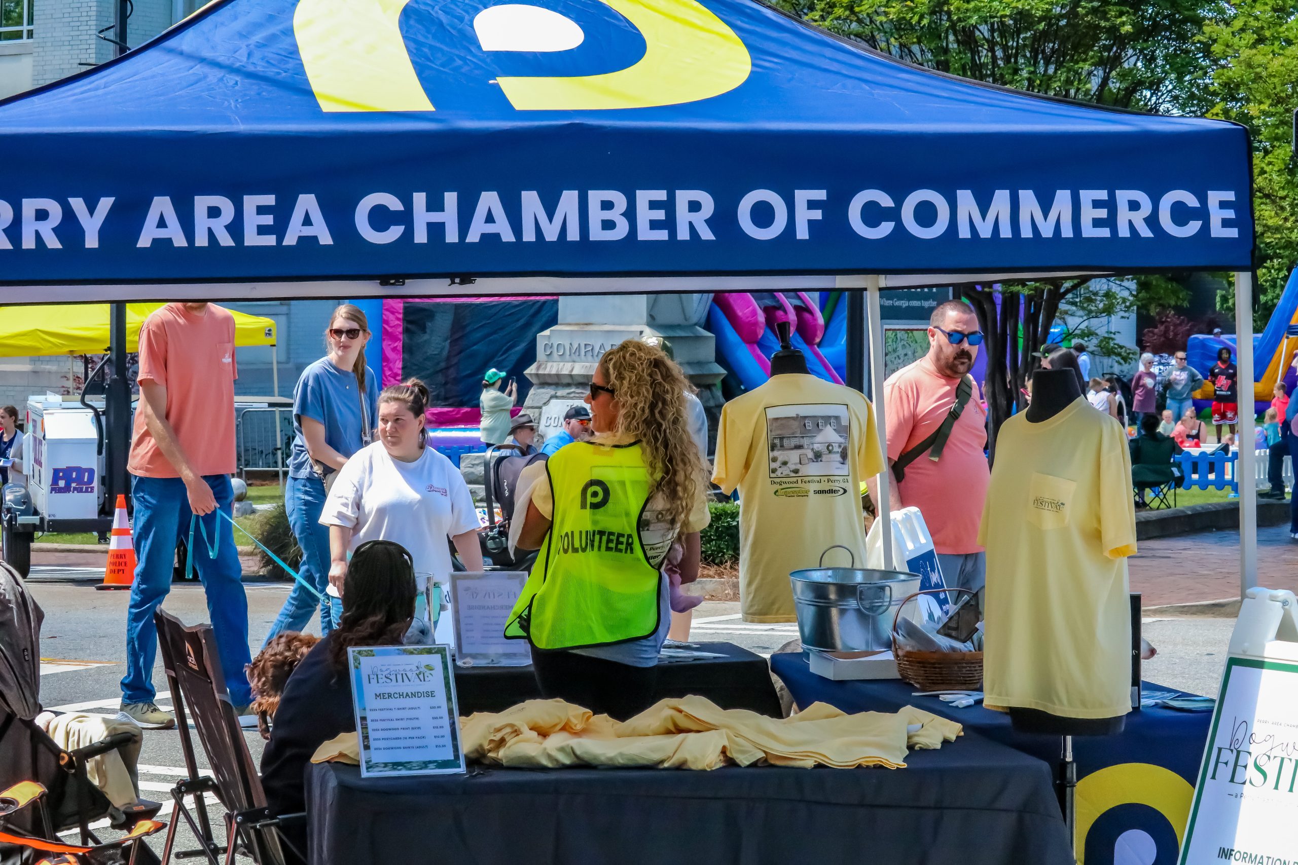 A Chamber of Commerce booth at an outdoor event displays yellow festival T-shirts and merchandise, with volunteers and visitors nearby.