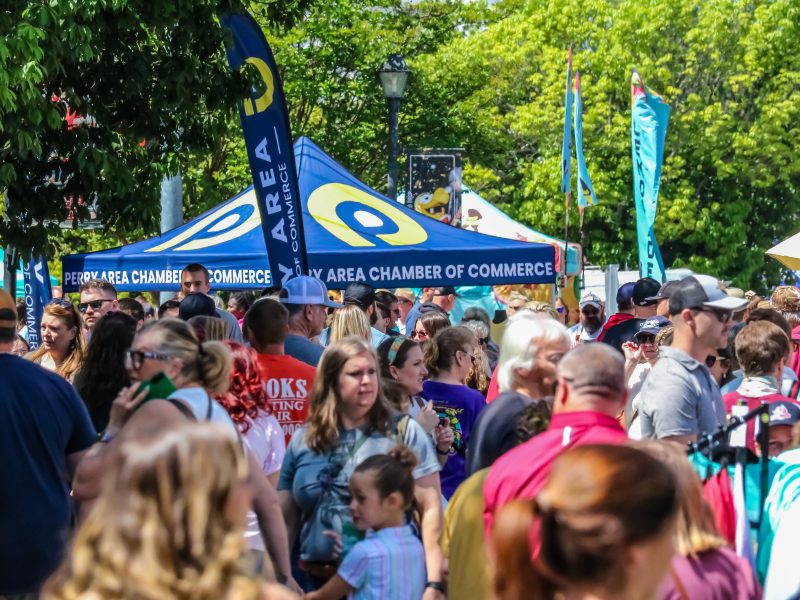 A large crowd walks through a sunny outdoor festival with Perry Area Chamber of Commerce tents and colorful flags.