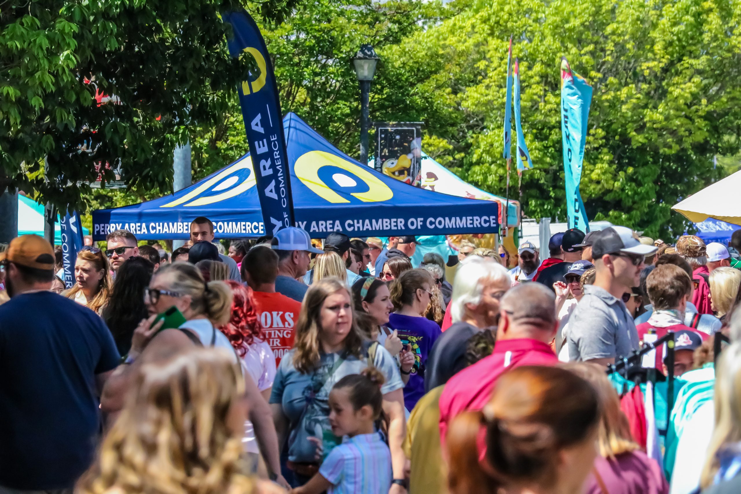 A large crowd walks through a sunny outdoor festival with Perry Area Chamber of Commerce tents and colorful flags.