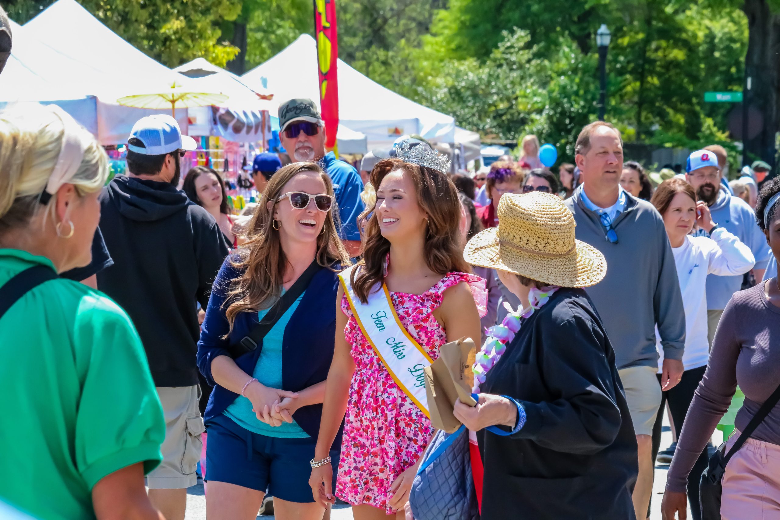 A young woman wearing a crown and “Teen Miss” sash smiles while walking through a crowded outdoor festival.