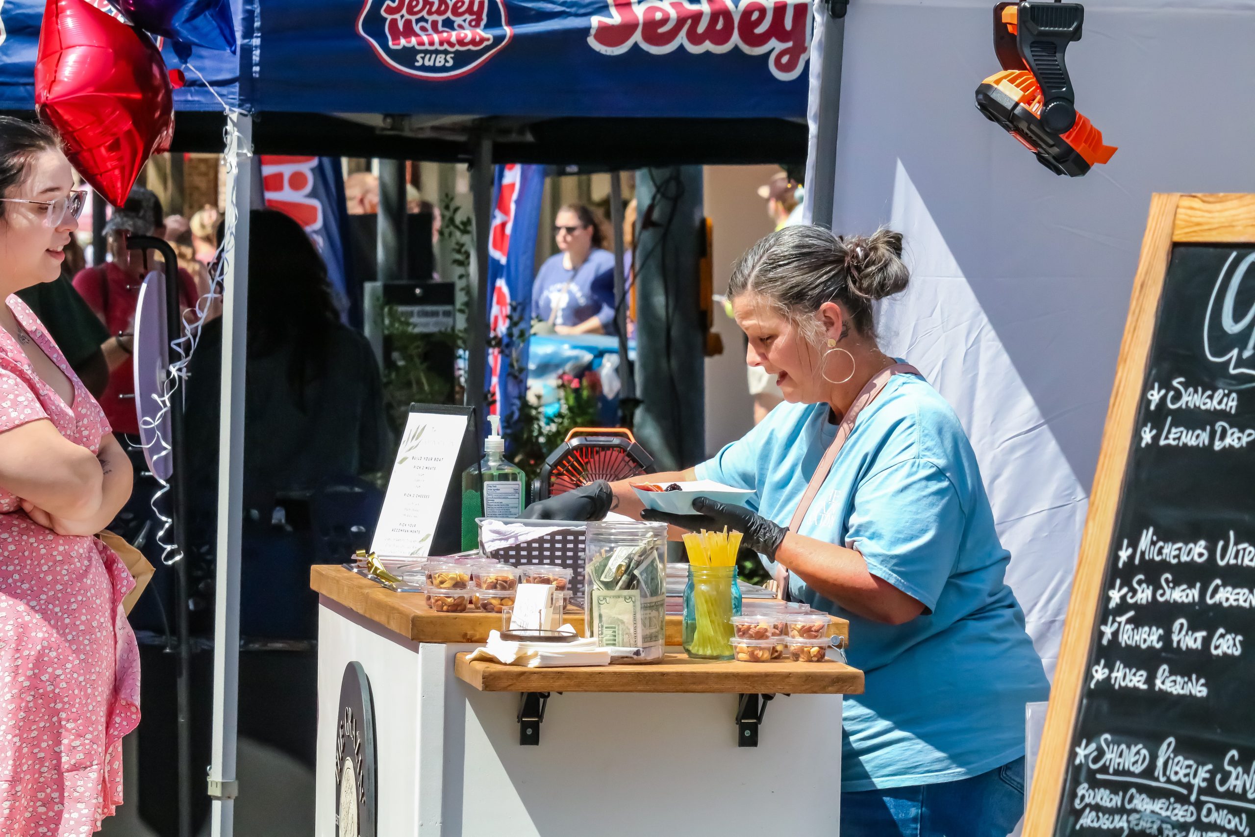 A vendor in a blue shirt serves food samples at an outdoor market booth, while a woman in a pink dress waits.