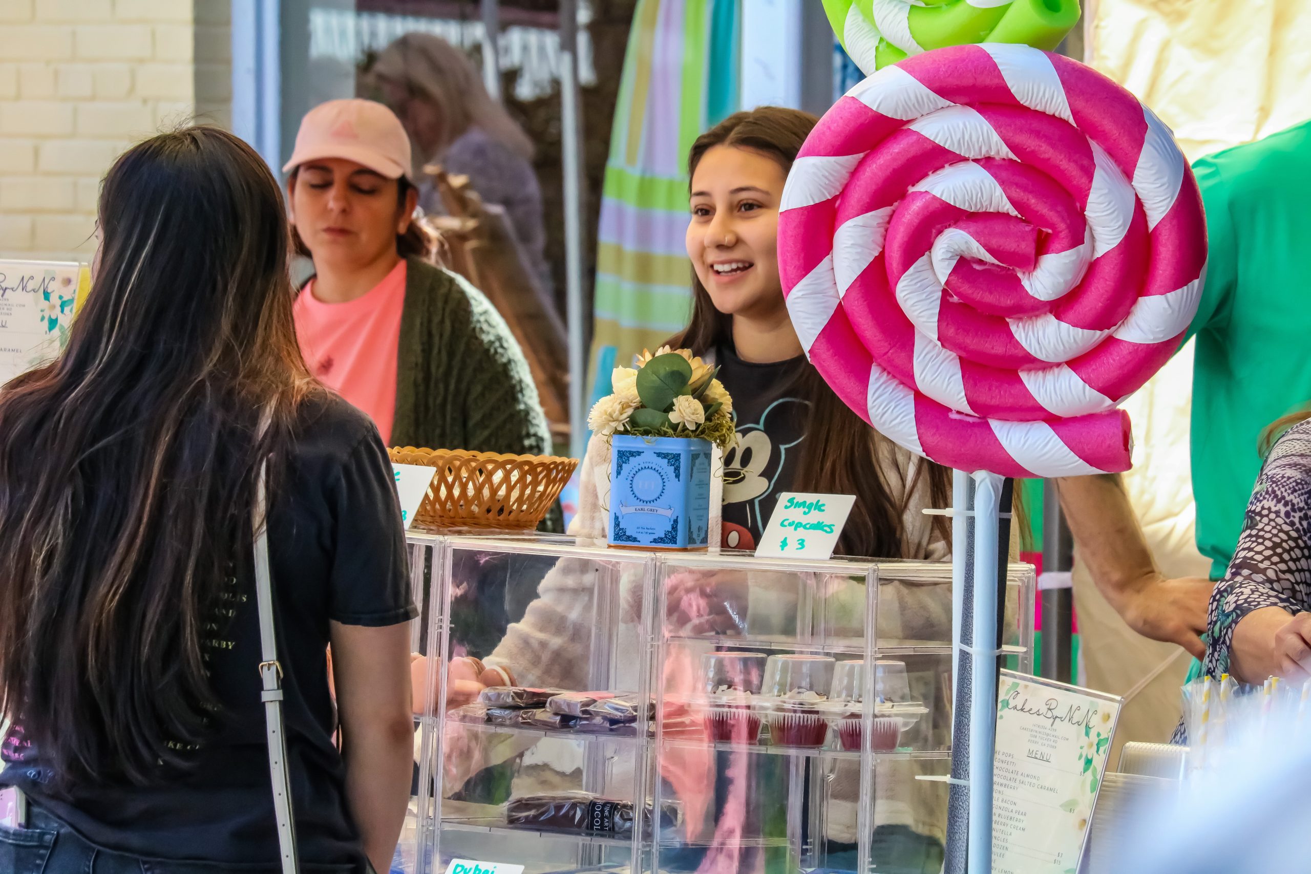 A young vendor smiles while selling cupcakes at a booth decorated with large candy props during an outdoor event.