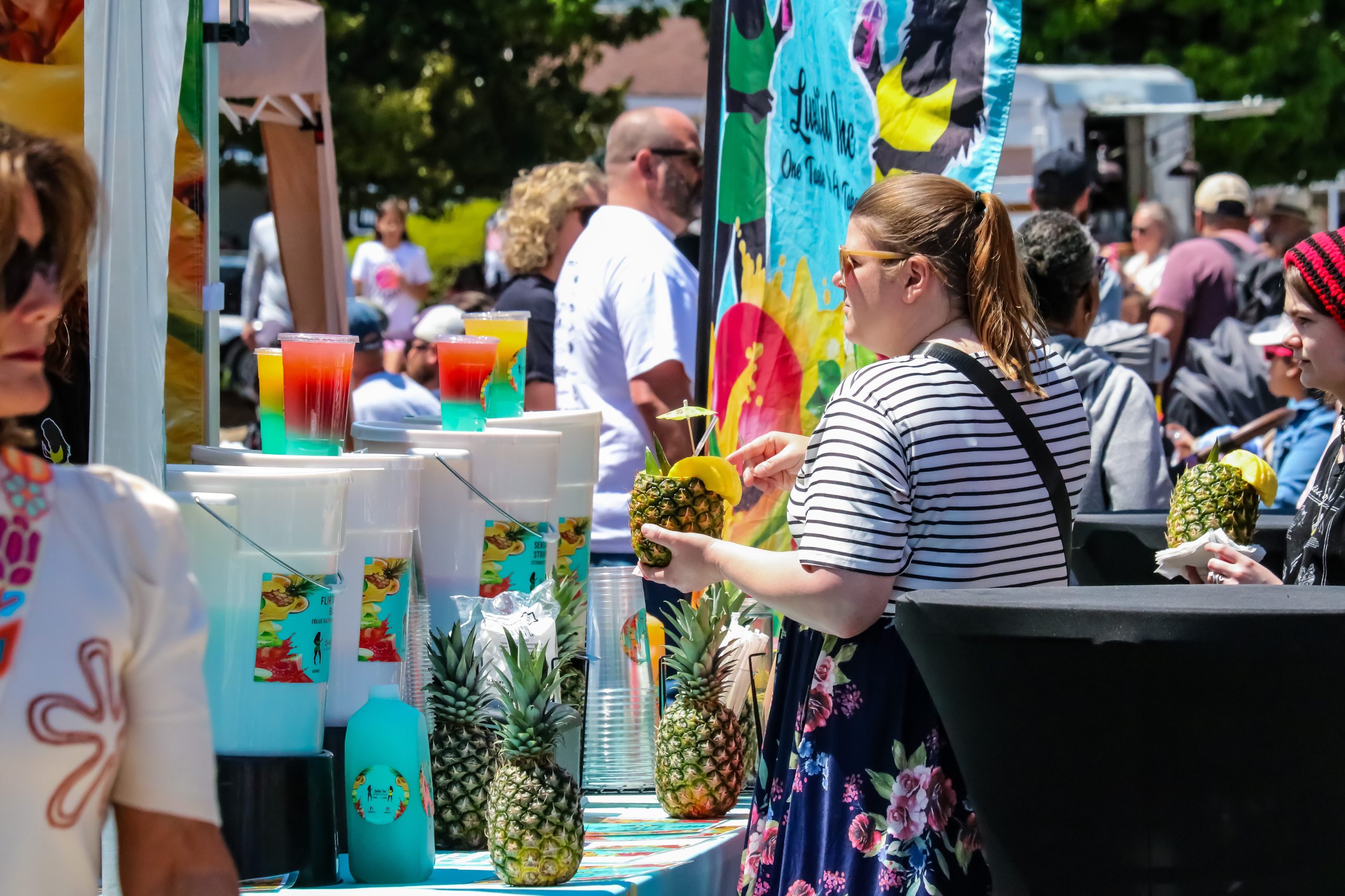A woman holds a tropical drink served in a pineapple at a colorful outdoor festival beverage stand.