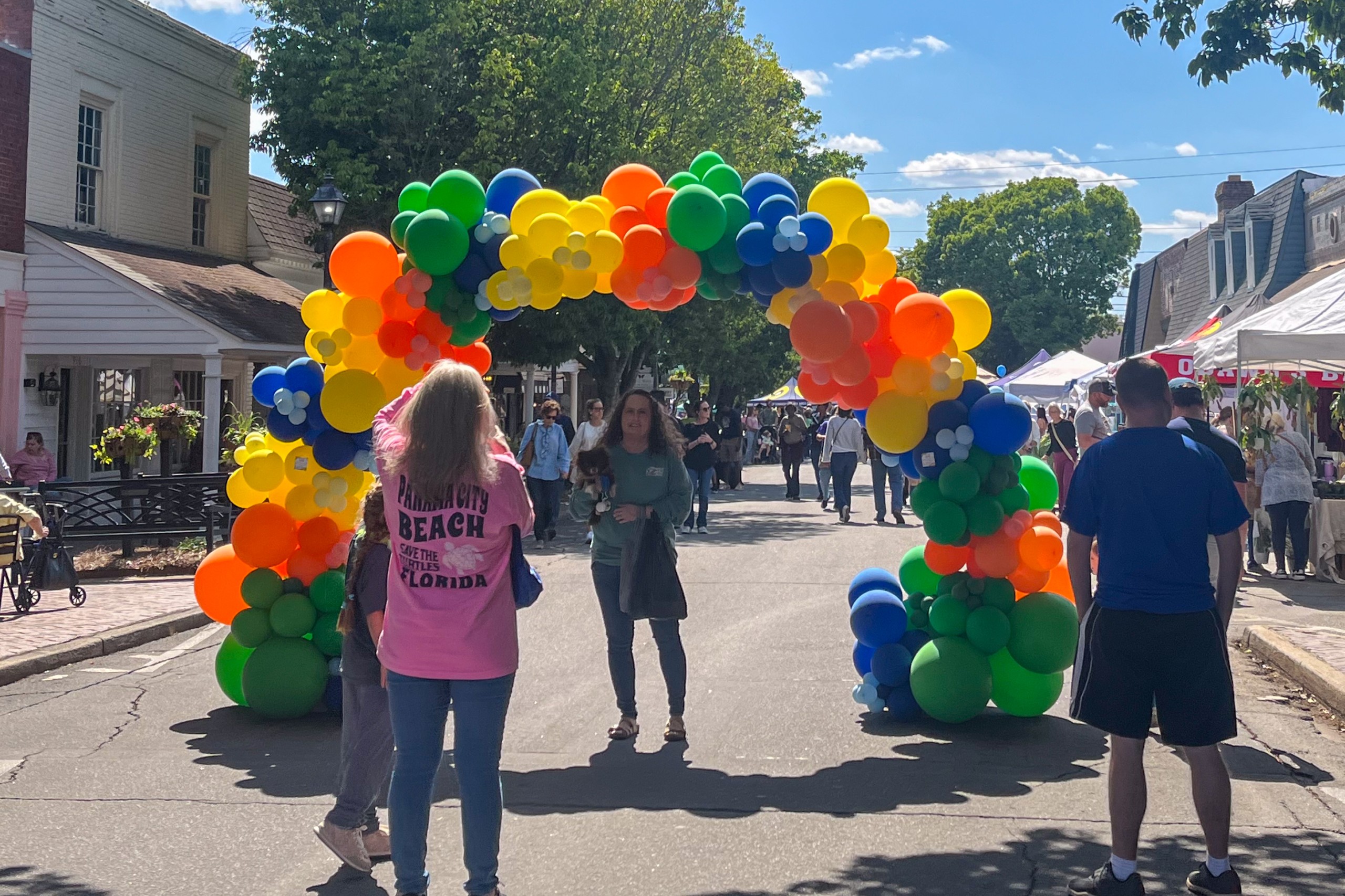 People walk under a colorful balloon arch at an outdoor street festival lined with vendor tents.