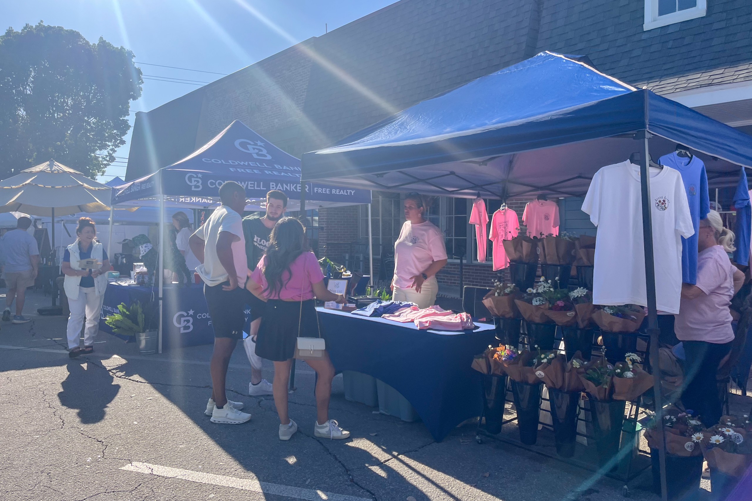 People visit vendor booths at an outdoor event, browsing flowers, T-shirts, and real estate information under blue tents.