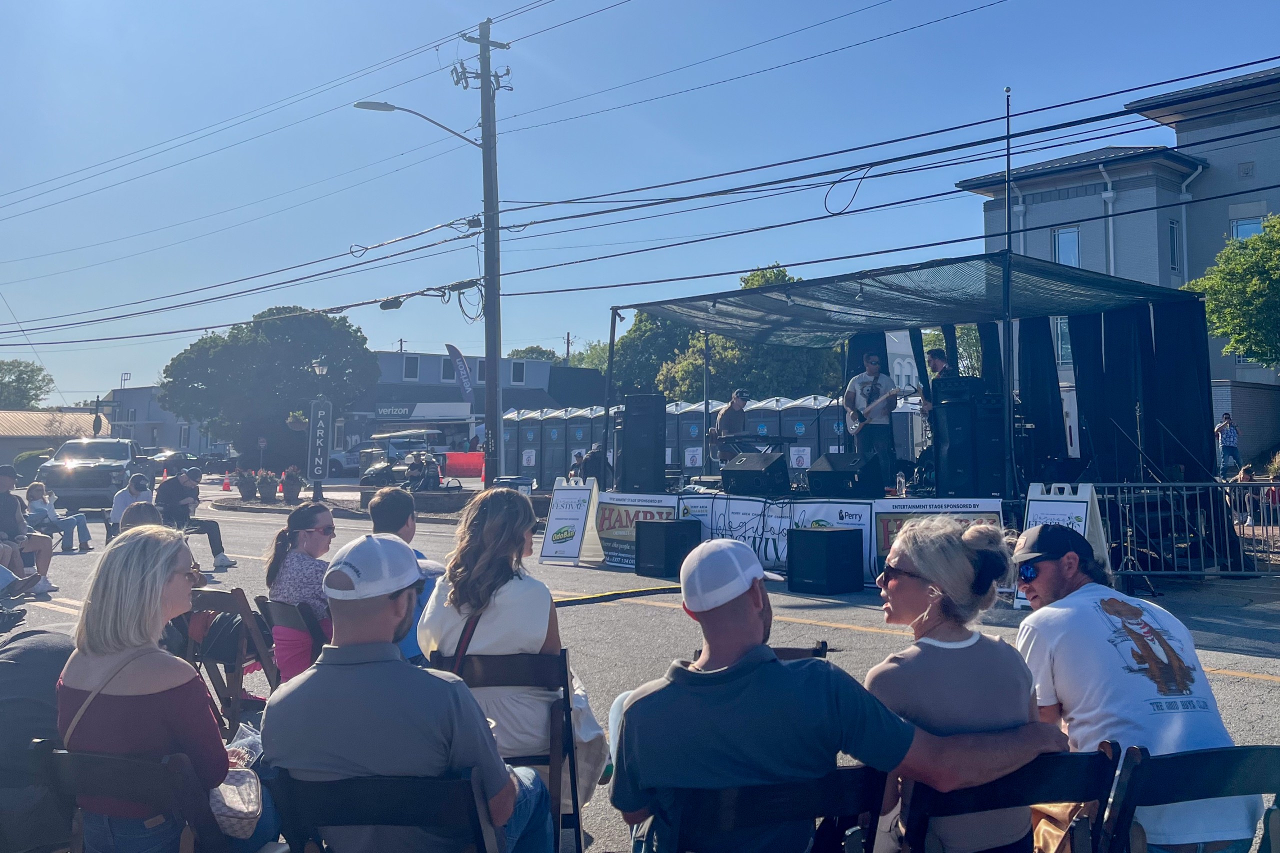 People sit in folding chairs watching a live band perform on an outdoor stage at a sunny street festival.
