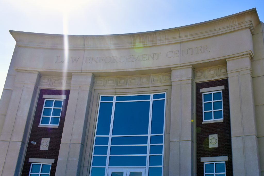 Exterior of a Law Enforcement Center with large glass windows, a curved beige facade, and sunlight streaming over the building’s name etched above the entrance. 