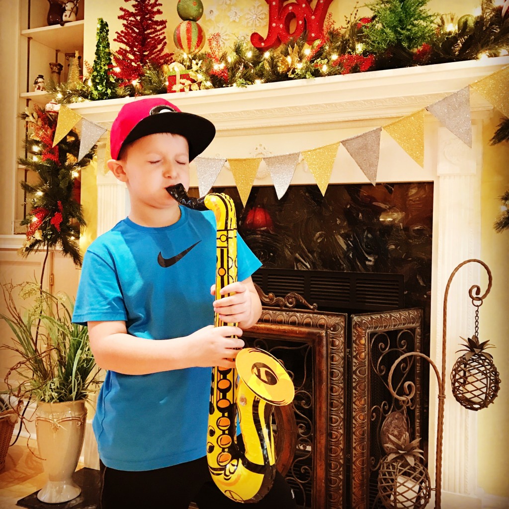 A little boy with a blue shirt and red hat playing the saxophone. Christmas decorations are seen in the background.