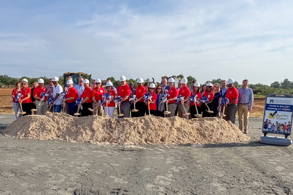 A group of people standing behind a symbolic pile of dirt for an outdoor groundbreaking ceremony, wearing hardhats and holding shovels.