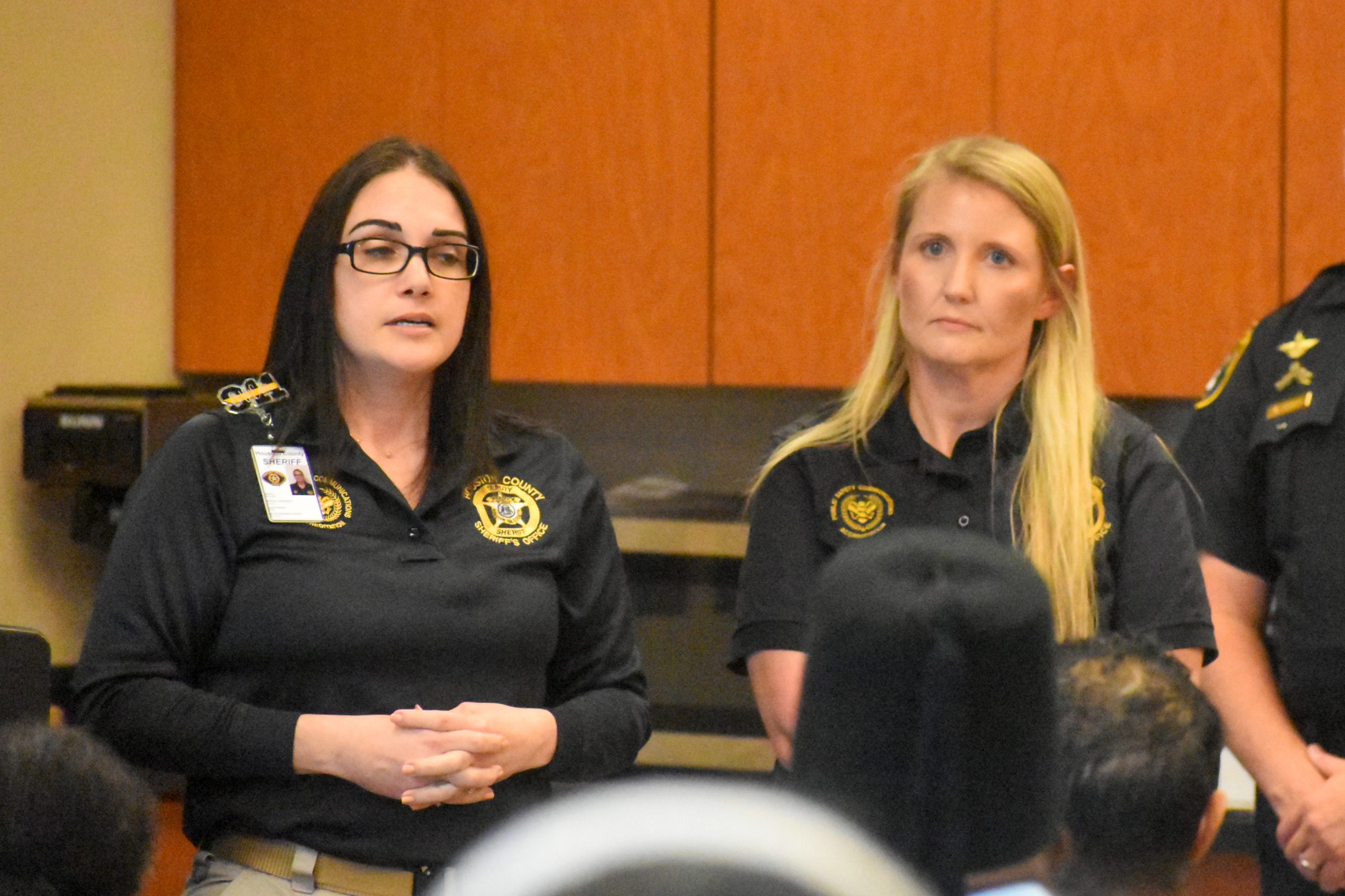 Two women in sheriff’s department uniforms stand and address an audience during an indoor event.