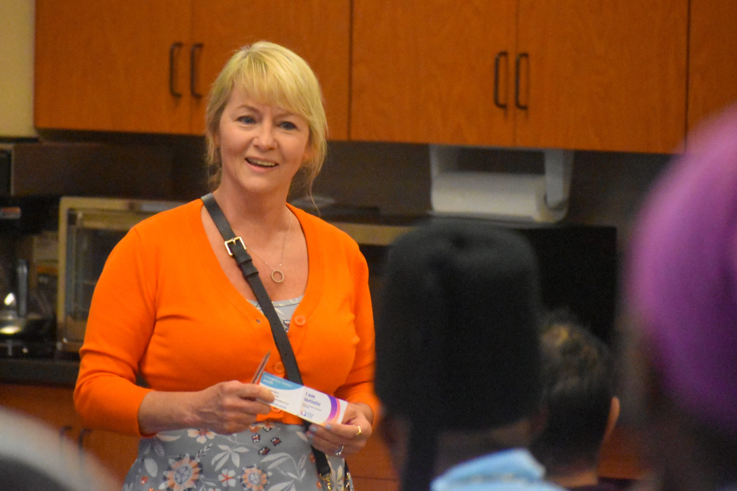 A smiling woman in an orange cardigan holds a small card while speaking to a seated audience.