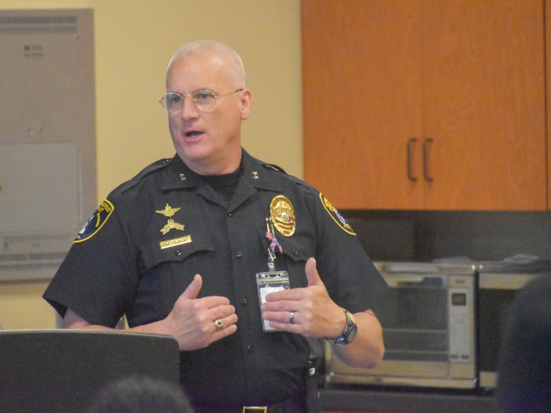 A uniformed police officer speaks while gesturing with his hands during an indoor presentation.