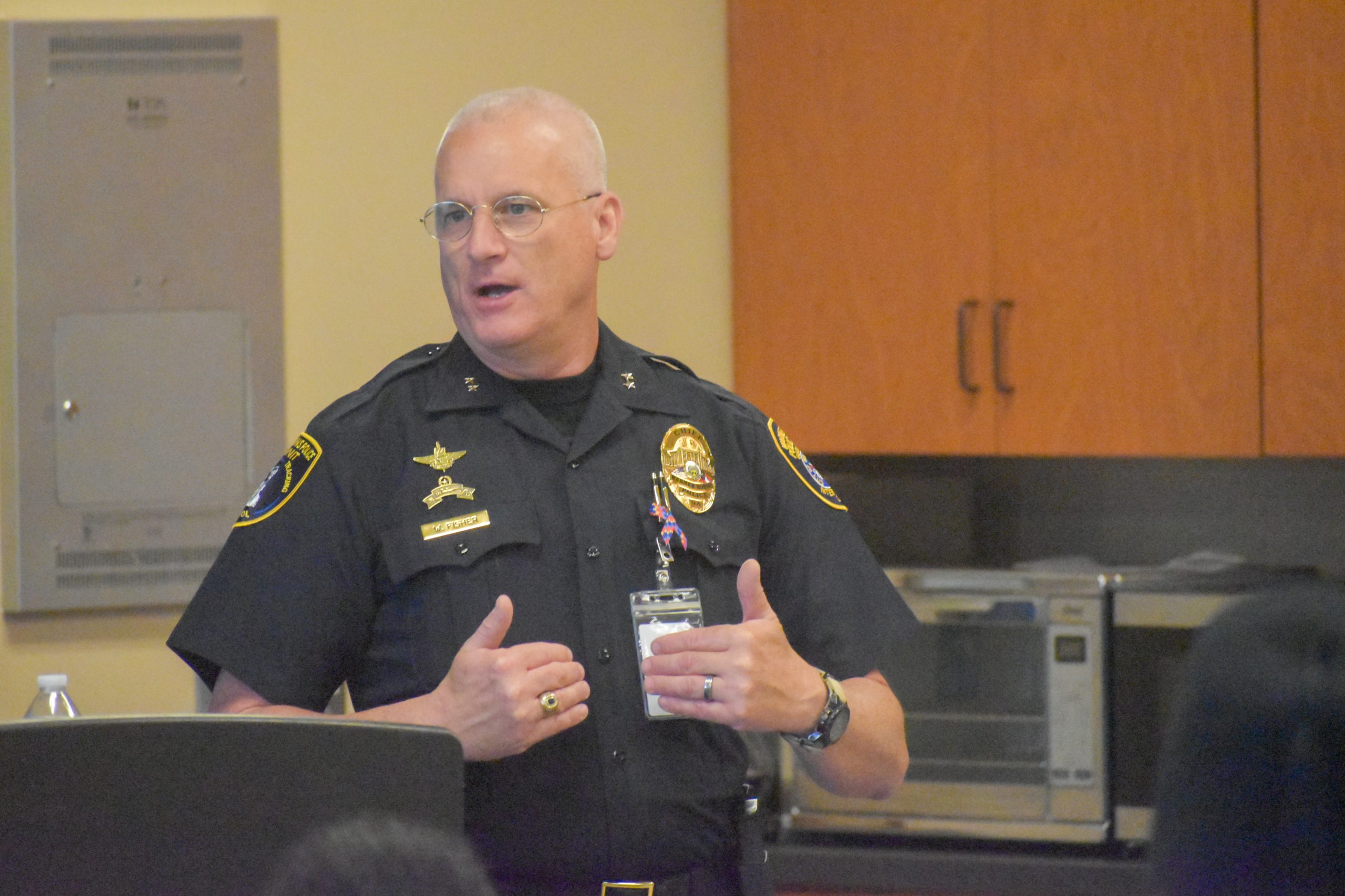 A uniformed police officer speaks while gesturing with his hands during an indoor presentation.
