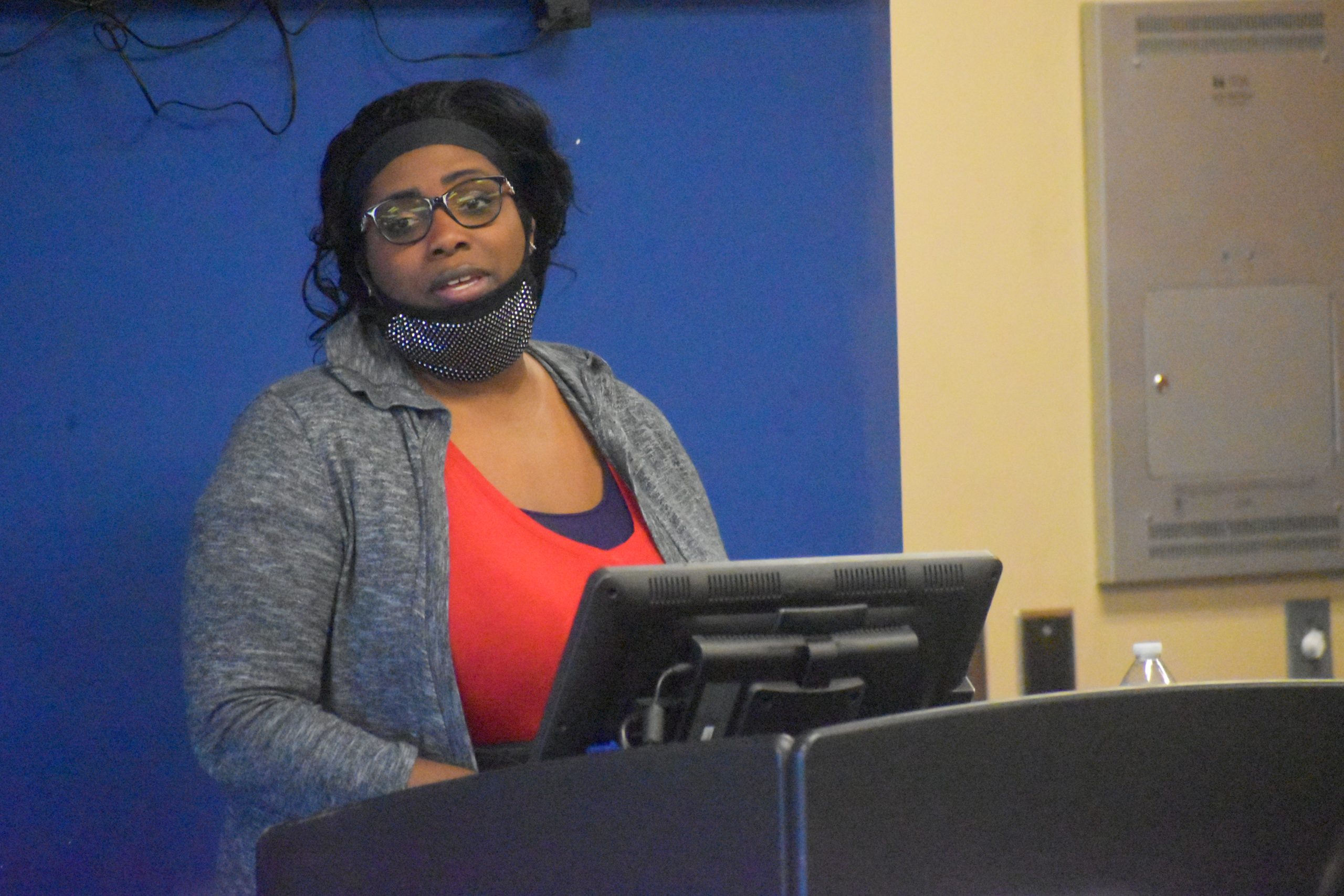 A woman wearing glasses and a face mask speaks at a podium in front of a blue wall.