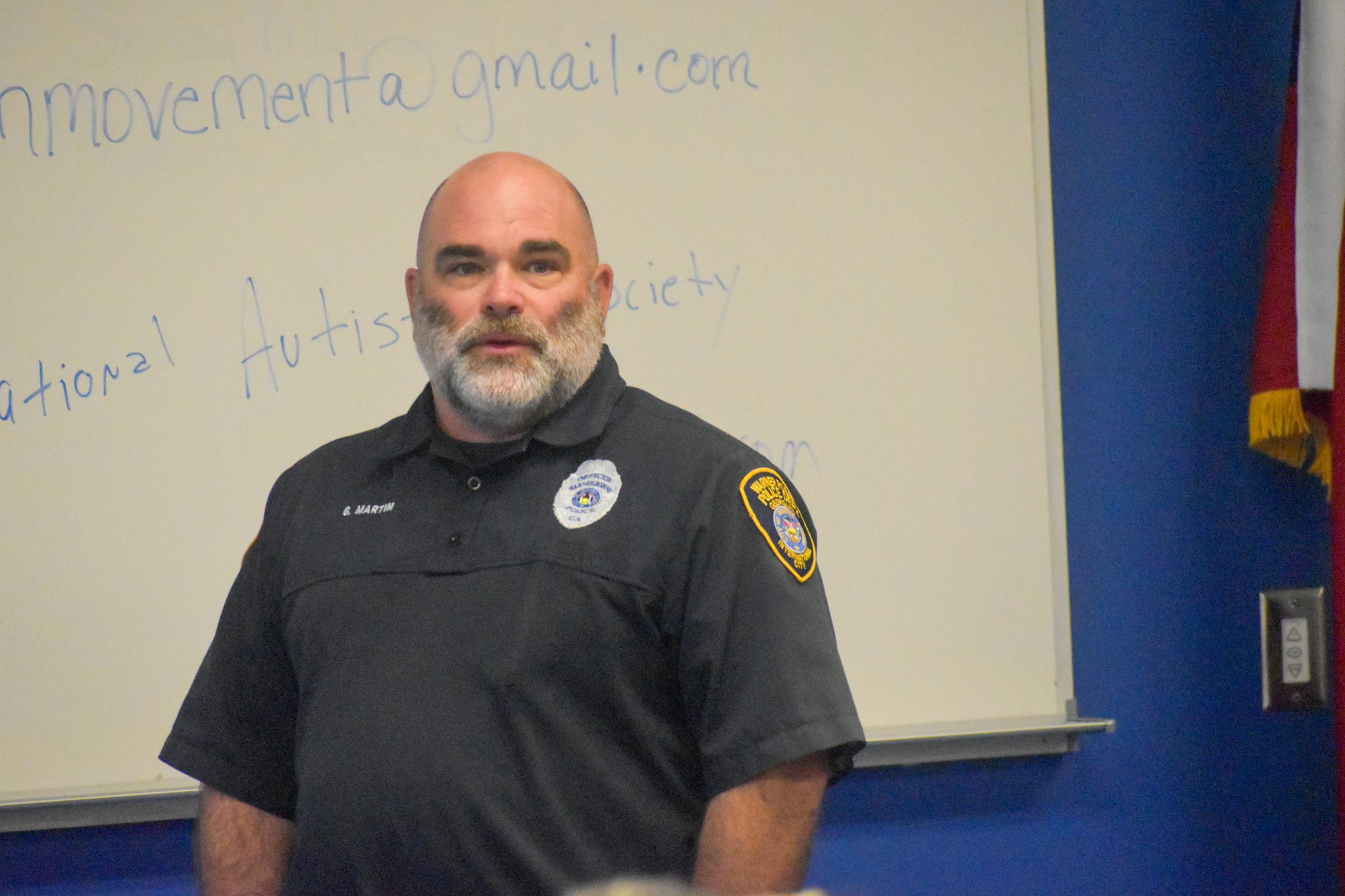 A uniformed officer stands in front of a whiteboard with handwritten text, addressing an audience.