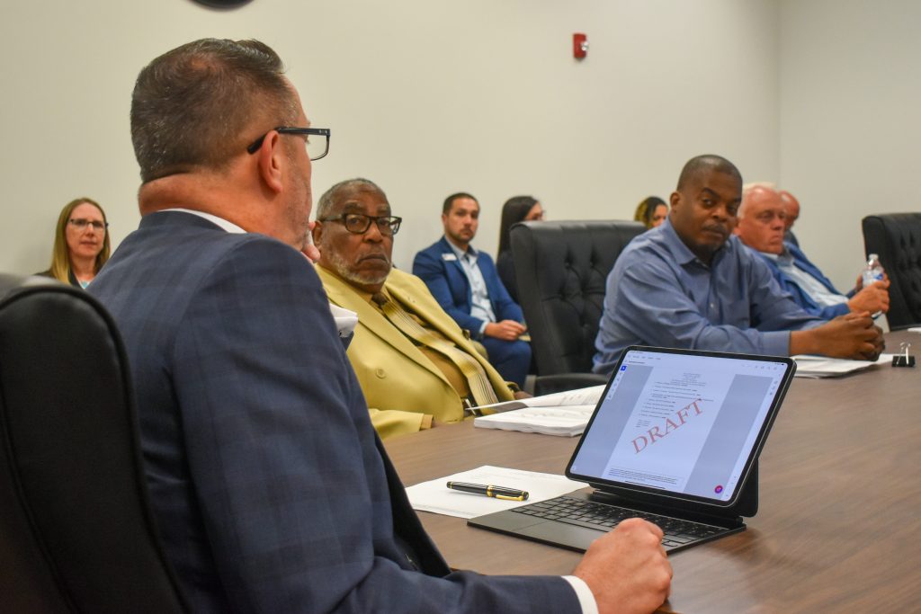 A group of people in a formal meeting room, with a man showing a digital document labeled “DRAFT” on a tablet screen.