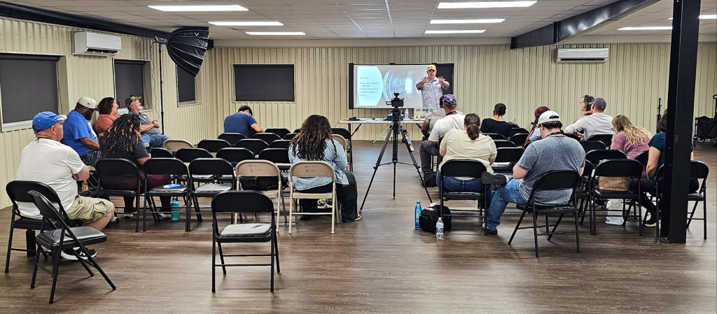 A room with people sitting down. A man is standing up giving a lesson.
