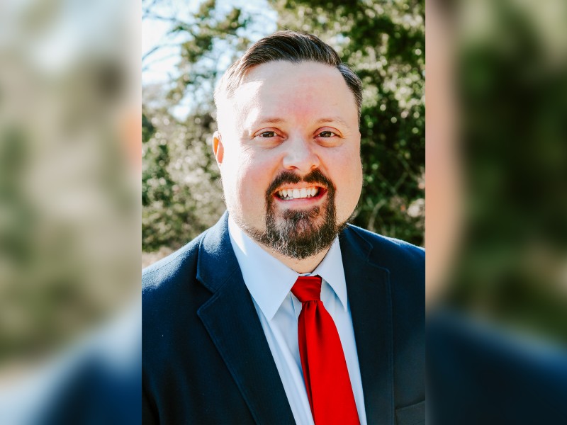 A man with a suit and a red tie. There is a blurry background.